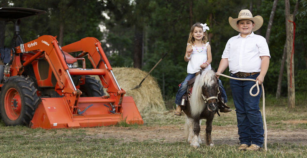 Tarver and Cecillia posing next to an orange tractor. Tarver is leading his sister, Cecilia, who is sitting on a pony. 